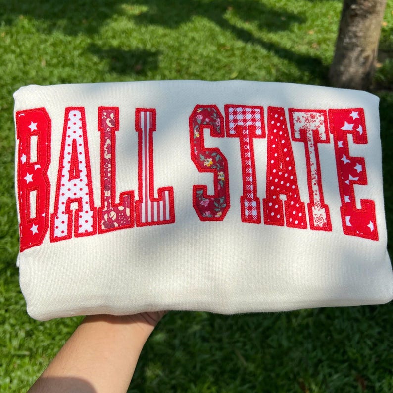 May include: A white sweatshirt featuring the text BALL STATE in large, block-style letters. Each letter is filled with a variety of red and white patterns, including floral prints, polka dots, gingham checks, and star motifs, with a red outline.