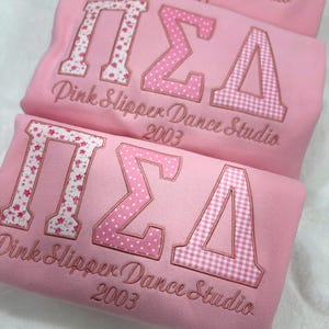 May include: Three folded light pink sweatshirts featuring large Greek letters appliquéd in floral, polka dot, and gingham patterns. Below the letters, the text Pink Slipper Dance Studio 2003 is embroidered in a dark pink script font.