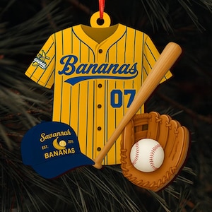 Pode incluir: Enfeite de Natal com tema de beisebol. Apresenta uma camisa amarela com riscas azuis e a palavra "Bananas". Inclui um bast&atilde;o de beisebol, uma luva, uma bola de beisebol e um bon&eacute; azul com o texto "Savannah Bananas". A camisa tem o n&uacute;mero "07".