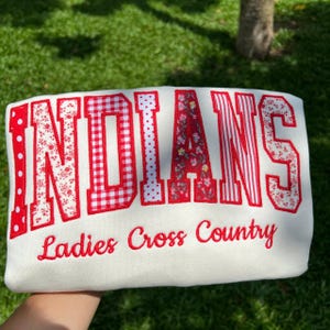 May include: A white sweatshirt featuring the word INDIANS in large, red-toned, patterned fabric letters, including polka dots, gingham, floral, and striped designs. Below the word, the text Ladies Cross Country is embroidered in red script.