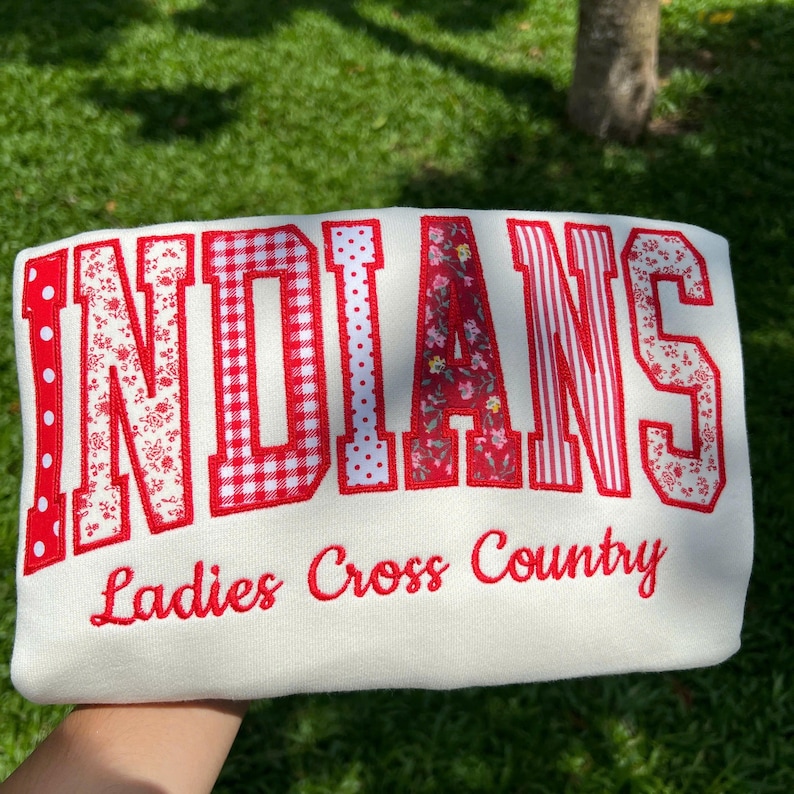 May include: A white sweatshirt featuring the word INDIANS in large, red-toned, patterned fabric letters, including polka dots, gingham, floral, and striped designs. Below the word, the text Ladies Cross Country is embroidered in red script.
