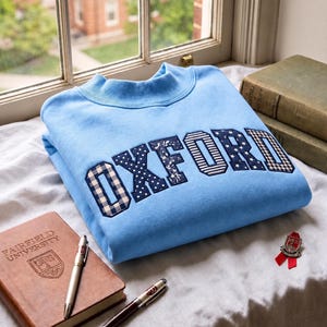 May include: A light blue crewneck sweatshirt featuring the word OXFORD appliqued across the chest in dark blue fabric with various patterns including gingham, polka dots, floral, and stars. The sweatshirt has a ribbed collar and long sleeves.