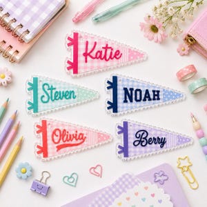 May include: Five triangular pennant-shaped acrylic keychains featuring names Katie, Steven, Noah, Olivia, and Berry. Each pennant has a gingham pattern, a solid color side strip, and a scalloped edge. The names are in raised, colorful script or block lettering.