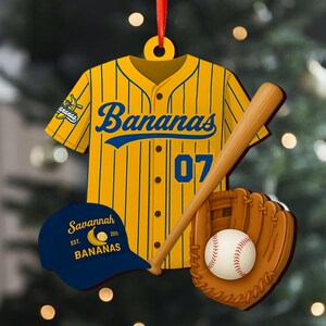 Pode incluir: Um enfeite com tema de beisebol com uma camisa amarela e azul com a palavra "Bananas" e o n&uacute;mero "07". Inclui um bast&atilde;o de beisebol, uma luva, uma bola e um bon&eacute; azul com o texto "Savannah Bananas". Uma fita vermelha est&aacute; presa.