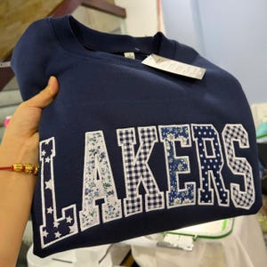 May include: A navy blue crewneck sweatshirt featuring the word LAKERS in large block letters. Each letter is filled with a different white and blue pattern, including stars, floral prints, gingham checks, and polka dots, with white stitching.