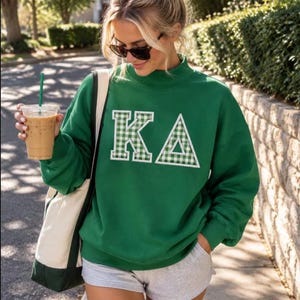 May include: A forest green mock neck sweatshirt featuring large white and green gingham patterned Greek letters KA on the chest. The sweatshirt has long sleeves and a relaxed fit, paired with light gray shorts and a canvas tote bag.