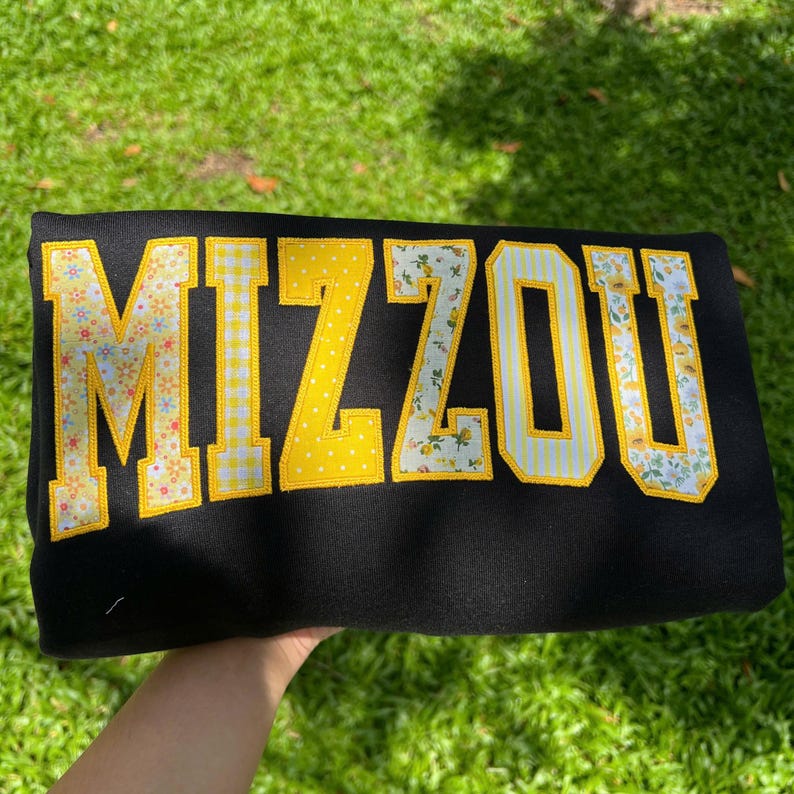 May include: A black sweatshirt featuring the word MIZZOU in large block letters. Each letter is appliqued with a different yellow and white fabric pattern, including floral prints, gingham checks, and polka dots, all outlined in yellow thread.