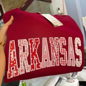 May include: A maroon crewneck sweatshirt featuring the word ARKANSAS in large, block-style applique letters. Each letter is crafted from different red and white patterned fabrics, including stars, floral prints, polka dots, and gingham checks.