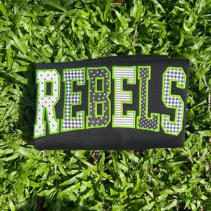 May include: A black fabric item featuring the word REBELS in large, block-style letters. Each letter is outlined in bright green and filled with various patterns, including white polka dots, black and white gingham, stars, and horizontal stripes.
