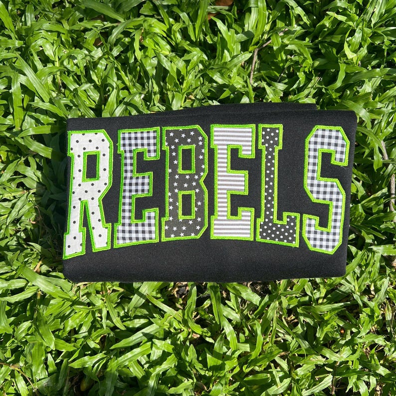 May include: A black fabric item featuring the word REBELS in large, block-style letters. Each letter is outlined in bright green and filled with various patterns, including white polka dots, black and white gingham, stars, and horizontal stripes.