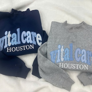 May include: A navy blue sweatshirt and a heather gray sweatshirt, each featuring the text "vital care" in a light blue patterned appliqué font with the word "HOUSTON" printed in white underneath the appliqué on the front of the garments.