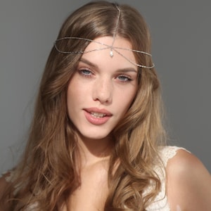 May include: A silver-toned headpiece with delicate chain details and a small, dangling pearl. The headpiece is worn across the forehead and over the hair, adding a touch of elegance. The design is simple yet eye-catching.