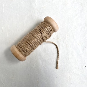 May include: A wooden spool wrapped with a length of natural tan jute twine. The spool features rounded ends and a smooth, light-colored wood finish. A single loose end of the brown fibrous cord extends from the main coil of the spool.