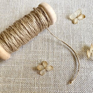 May include: A wooden spool wrapped with natural twine, lying on a textured beige surface. Dried, tan-colored flower petals are scattered around the spool. The twine extends from the spool, suggesting it's ready for use.
