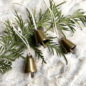 May include: Three small, antique brass bells with white string hangers are displayed on a bed of green cedar branches and a fluffy white surface. The bells have a classic, conical shape and are ideal for holiday decor.