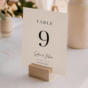 May include: A cream-colored table number card with the number 9 in a bold, black font. The card also includes the names "Sofia & Julian" and the date "10 MAY 2027". The card is held in a light wood block. Wedding decor.
