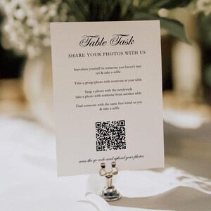 Puede incluir: Una tarjeta blanca con el texto "Table Task" y "Share Your Photos With Us" está sujeta por un soporte de metal plateado. La tarjeta incluye instrucciones y un código QR para compartir fotos en una boda o evento.