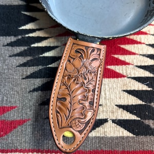Puede incluir: Una funda de cuero marrón para el mango de la sartén con un diseño en relieve de hojas de roble y bellotas. La funda está cosida alrededor de los bordes y tiene un agujero para colgar. La funda está junto a una sartén gris sobre una alfombra estampada.
