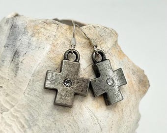 May include: A pair of silver-toned cross earrings with a small, clear gem in the center of each cross. The earrings hang from silver-colored hooks. The earrings are displayed on a light-colored, textured surface.