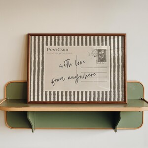 May include: A rectangular framed print featuring a postcard design with the text "with love from anywhere" in cursive script, surrounded by a dark gray and white vertical striped border, resting on a sage green wall-mounted shelf with light trim.