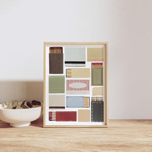 May include: A framed collage of matchboxes with various colors and patterns, including red, white, blue, green, and brown. The matchboxes are arranged in a grid pattern and are displayed against a white background.