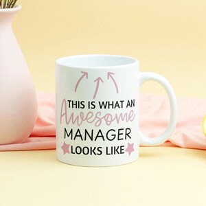 Può includere: Una tazza in ceramica bianca con un design rosa che dice "This is what an Awesome Manager looks like" con due stelle su entrambi i lati del testo. La tazza è appoggiata su una superficie rosa e bianca.