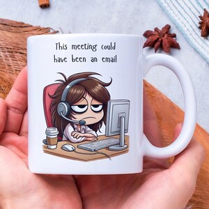 May include: White ceramic mug featuring a cartoon illustration of a person wearing headphones and looking weary at a computer. The text on the mug reads "This meeting could have been an email."