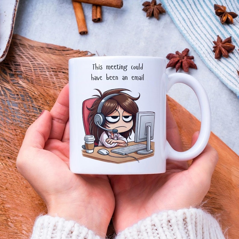 May include: White ceramic mug featuring a cartoon illustration of a person wearing headphones and looking weary at a computer. The text on the mug reads "This meeting could have been an email."