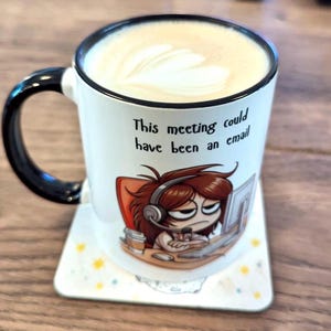 May include: White ceramic mug with a black handle and a cartoon illustration of a person wearing headphones and looking annoyed at a computer screen. The mug has the text "This meeting could have been an email" printed on it.