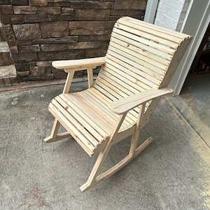 May include: A wooden rocking chair with a slatted back and seat, and armrests. The chair is made of light-colored wood and has a natural finish. The chair is designed for outdoor use and is placed on a concrete surface.