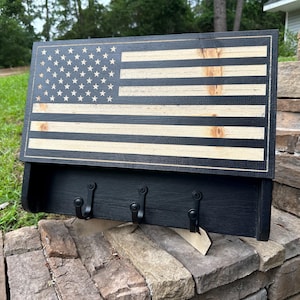 May include: A wooden wall-mounted key holder and shelf, featuring the American flag design. The flag is crafted with alternating black and natural wood stripes and stars. The shelf has three black metal hooks for keys.