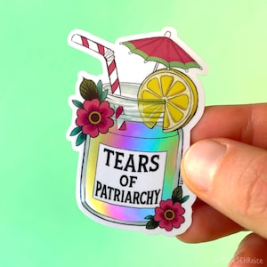 Könnte beinhalten: Ein holografischer Aufkleber mit einem Cocktail mit einer Zitronenscheibe, einem roten Sonnenschirm und einem gestreiften Strohhalm. Das Glas ist mit rosa Blumen und dem Text "TEARS OF PATRIARCHY" verziert.