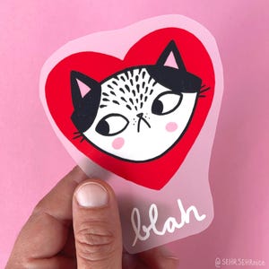 Könnte beinhalten: Ein Aufkleber mit einem Katzenkopf in einem roten Herz. Die Katze hat schwarz-weiße Markierungen und einen mürrischen Gesichtsausdruck. Das Wort "blah" ist in weißer Schrift unter dem Herzen geschrieben. Der Aufkleber wird vor einem rosa Hintergrund gehalten.