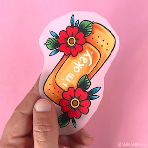 Könnte beinhalten: Ein Aufkleber mit einem orangefarbenen Pflaster-Design mit schwarzen Punkten und roten Blumen mit grünen Blättern. Der Text "I'm okay" ist auf dem Pflaster geschrieben. Der Aufkleber wird vor einem rosa Hintergrund gehalten.