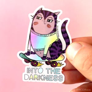 Könnte beinhalten: Ein Aufkleber mit einer Cartoon-Katze mit lila und weißen Streifen, die einen Regenbogenpullover trägt und auf einem Skateboard sitzt. Der Aufkleber trägt den weißen Text "INTO THE DARKNESS". Der Hintergrund ist hellrosa.