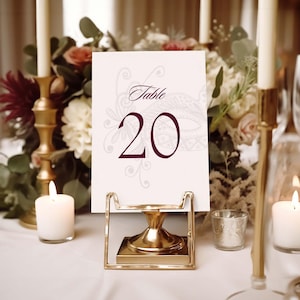 May include: A table number card with the number 20 in burgundy script, displayed in a gold metal holder. The card has decorative floral elements. The scene includes candles, flowers, and a gold candlestick.
