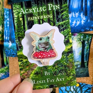 May include: An acrylic pin featuring a fairy frog perched on a red mushroom with white spots. The pin is on a card with the text "Acrylic Pin Fairy Frog" and "By Linzi Fay Art". The background shows a forest scene.
