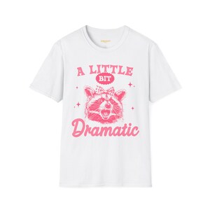 Puede incluir: Camiseta blanca con un gráfico rosa de un mapache con un lazo y el texto "A Little Bit Dramatic". El mapache está sonriendo. El texto está en una fuente juguetona.