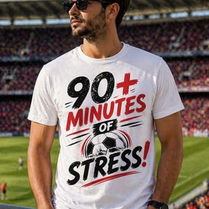 Puede incluir: Camiseta blanca con el texto "90+ MINUTES OF STRESS!" en negro y rojo, con un gráfico de balón de fútbol. La camiseta la lleva una persona con gafas de sol, de pie frente a un estadio.