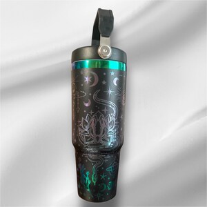 May include: Black insulated tumbler with a handle, featuring a celestial design with a lotus flower, snake, stars, and moons. The top rim has a teal and green iridescent band. The handle is black and attached with a silver clasp.
