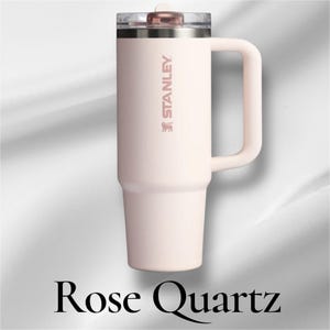 K&ouml;nnte beinhalten: Ein blassrosa Stanley-Becher mit passendem Seitengriff und einem transparenten Deckel mit rotierendem Verschluss in Ros&eacute;gold. Der Markenname Stanley ist vertikal in einem metallischen Ros&eacute;goldton an der Seite des Isolierbechers aufgedruckt.