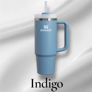K&ouml;nnte beinhalten: Ein matter blauer Stanley-Becher mit gro&szlig;em Seitengriff, wei&szlig;em Deckel und passendem wei&szlig;em Strohhalm. Das Markenlogo ist in Wei&szlig; auf der oberen Vorderseite aufgedruckt. Das Wort Indigo steht in einer schwarzen Serifenschrift unter dem Becher.