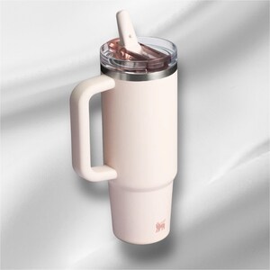 K&ouml;nnte beinhalten: Ein blassrosa isolierter Becher mit einem gro&szlig;en seitlichen Griff, einem durchsichtigen Kunststoffdeckel und einem passenden blassrosa Strohhalm. Der zylindrische Beh&auml;lter hat eine konische Basis und ein kleines Logo unten.