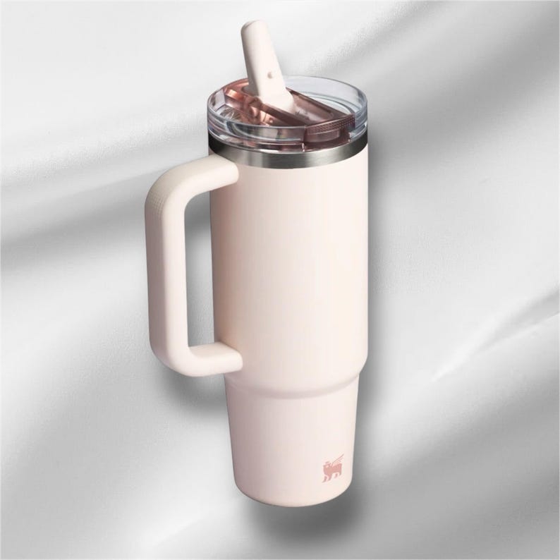 K&ouml;nnte beinhalten: Ein blassrosa isolierter Becher mit einem gro&szlig;en seitlichen Griff, einem durchsichtigen Kunststoffdeckel und einem passenden blassrosa Strohhalm. Der zylindrische Beh&auml;lter hat eine konische Basis und ein kleines Logo unten.