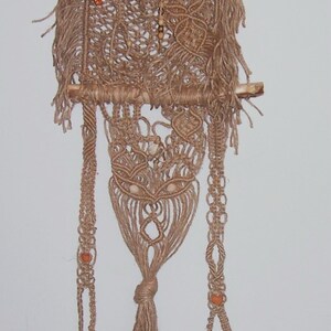 May include: A macrame wall hanging with a brown and beige color scheme. The hanging features a woven design with beads and shells. The hanging is attached to two wooden dowels.