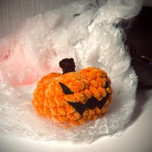 Peut inclure: Une citrouille orange en crochet avec un sourire noir et dentu. La citrouille est posée sur une surface blanche et moelleuse.