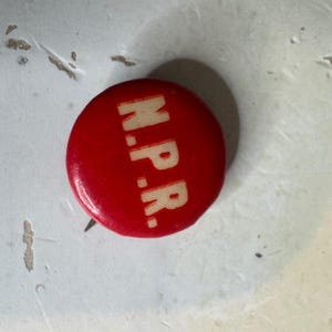 Op de afbeelding: Een ronde rode pin-back button met de witte tekst N.P.R. in een vet, schreefloos lettertype in het midden. De button heeft een gladde, glanzende afwerking en een standaard metalen speldmechanisme dat zichtbaar is aan de achterkant van het object.