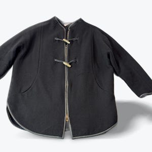 May include: Black jacket with a unique design. The jacket features a zipper closure and toggle closures. The jacket has a rounded hemline and long sleeves. The jacket has a gray trim around the edges and pockets.
