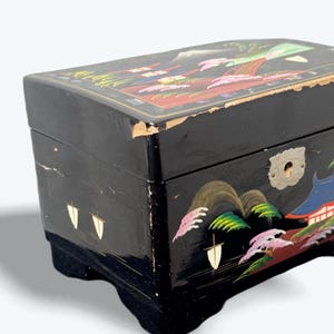 May include: Black lacquered jewelry box with a painted landscape design. The box features a keyhole and decorative metal hardware. The landscape design includes mountains, trees, and buildings in shades of red, green, and blue.