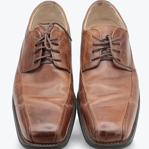 May include: A pair of brown leather dress shoes with a square toe design. The shoes have a lace-up closure and a dark brown sole. The leather has a slightly distressed appearance, suggesting regular wear. The shoes are set against a plain white background.